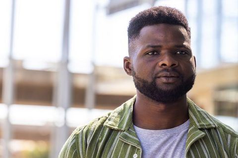 Thoughtful urban man standing in sunlit plaza wearing green striped overshirt and gray tee