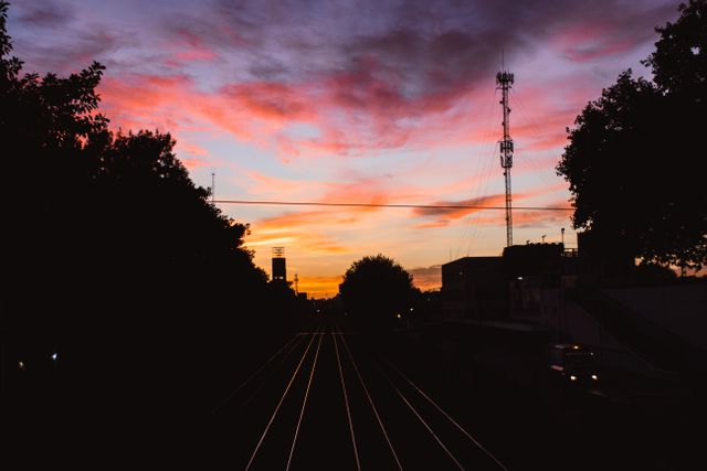 Twilight scene depicting sunset over railway tracks with silhouetted trees and radio tower against a colorful evening sky. Ideal for use in travel, transportation, nature, and cityscape themes, as well as background images for websites, presentations, and social media.