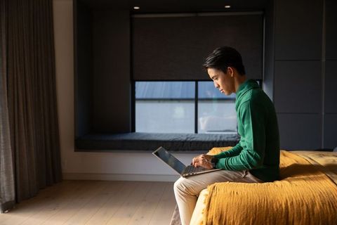 Asian man working on laptop on bed edge in modern minimalist bedroom with mustard throw