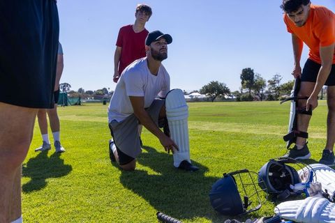 Cricket teammates fastening batting pads during sunny practice on grassy outdoor field