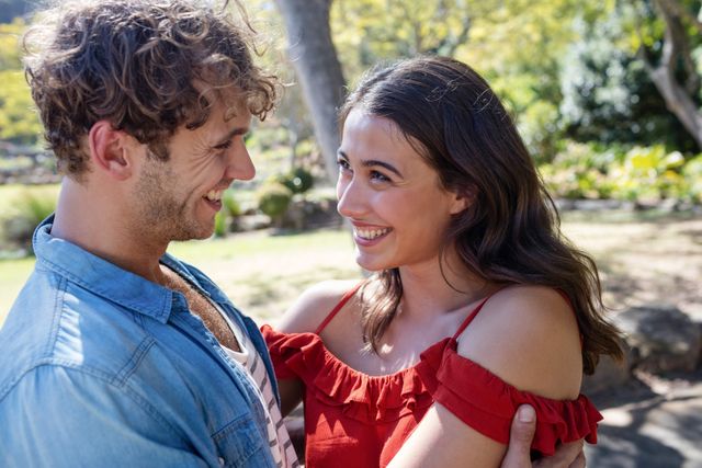 Romantic couple smiling at each other in a park on a sunny day. Ideal for use in advertisements, relationship blogs, social media posts about love and happiness, and lifestyle magazines.