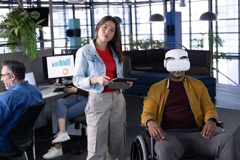 Wheelchair user experiencing vr while colleague guiding on tablet in modern office