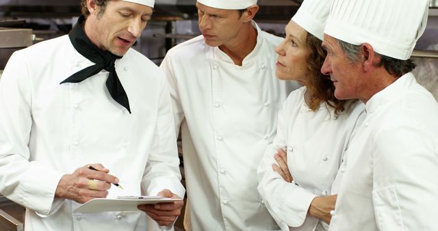 Chefs in uniform are gathered around, actively engaging in discussions about recipes or kitchen tasks. High-level collaboration is observed, making this useful for themes involving teamwork, professional kitchens, culinary arts, hospitality industry, and chef training programs.