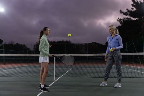 Female tennis coach training player at dusk on outdoor hardcourt during coaching drill