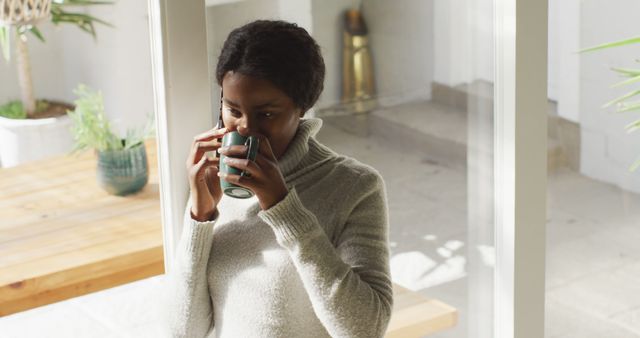 Young woman in cozy sweater sipping coffee while talking on smartphone. Ideal for use in lifestyle blog articles, advertisements for home comfort products, or promotional content for coffee and tea brands.