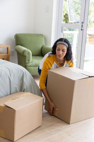 Young woman lifting moving box in bright bedroom during unpacking and moving day