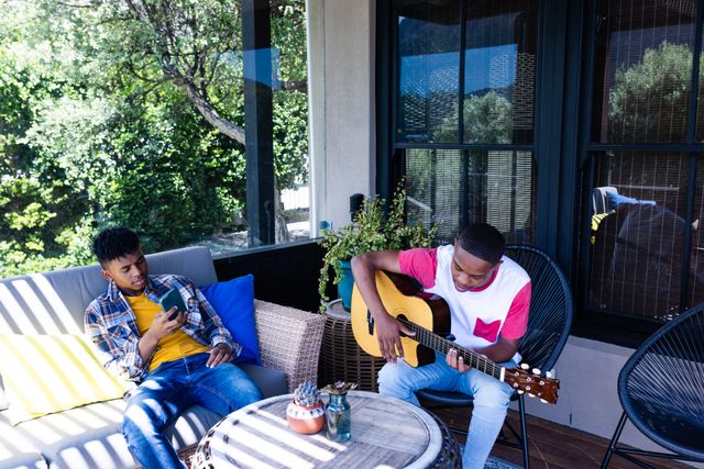 African american teenage male friends sitting on patio with smartphone and guitar. Hanging out with friends and spending quality time together concept.