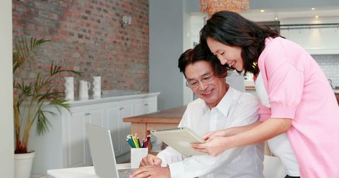 two asian adults collaborate on a digital project in a stylish home office setting, showcasing effective use of modern technology, like a laptop and a tablet. ideal for topics such as remote work solutions, teamwork, professional collaboration, and home office inspiration, this scene emphasizes digital literacy and innovation in professional practices.