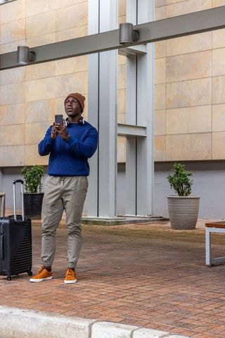 Urban traveler checking smartphone next to wheeled suitcase at modern building entrance