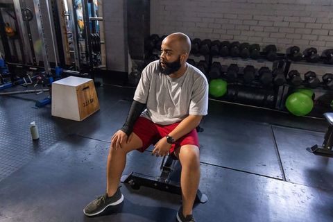 African american man sitting on bench wearing grey tee and red shorts during gym rest