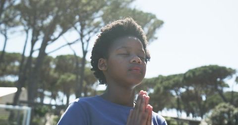 African teen meditating outdoors with hands clasped and eyes closed in sunlit pine park