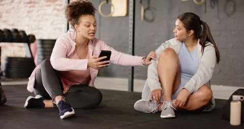 Female gym partners resting and chatting while tying sneakers and checking workout app