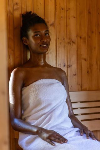 African american woman relaxing in wooden sauna wrapped in white towel for spa wellness