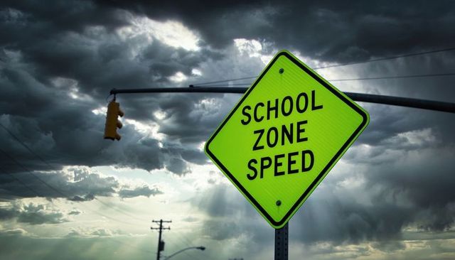 Fluorescent school zone speed sign tilting against storm clouds with traffic light