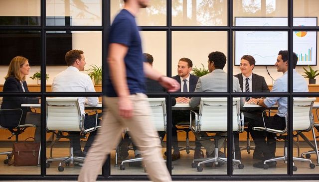 Executives discussing strategy in glass-walled conference room with passerby walking