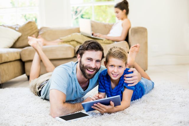 Father and son lying on a rug in the living room, using a digital tablet together. The father is smiling and has his arm around his son, who is also smiling. In the background, a woman is sitting on a couch using a laptop. This image can be used for family-oriented content, technology in family life, parenting articles, or advertisements promoting digital devices.