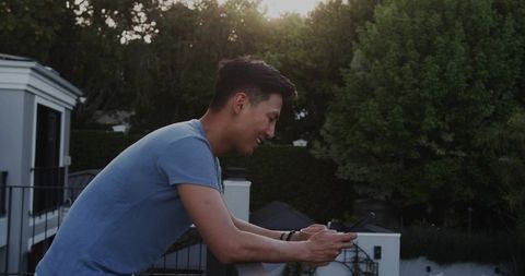 Young Asian man leaning on balcony railing checking smartphone during sunset