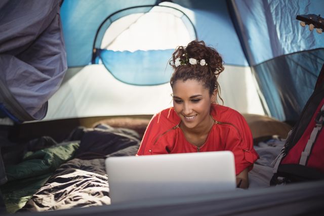 Young woman lying in a tent, smiling while using a laptop. Ideal for themes related to camping, outdoor activities, technology in nature, remote work, travel, and leisure. Suitable for promoting camping gear, travel blogs, digital nomad lifestyle, and outdoor adventure brands.