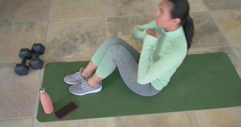 Mid-adult asian woman doing crunches at home on green mat with dumbbells and water bottle
