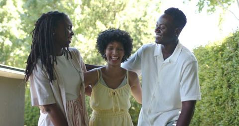 Friends laughing and leaning on sunlit porch railing by green garden, candid summer moment