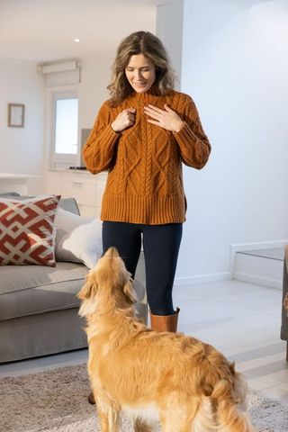 Mid-adult woman adjusting rust sweater while greeting golden retriever in cozy living room