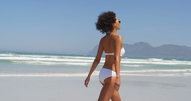 This image captures a young woman walking along a sunny beach, wearing a white bikini and sunglasses. The clear blue sky and gentle waves in the background evoke a sense of relaxation and escape. Ideal for use in travel blogs, vacation promotions, beachwear advertisements, posters, and lifestyle articles.