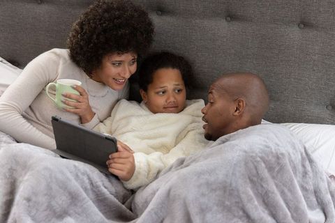 African american family cuddling in bed with tablet and mug, cozy morning bonding moment