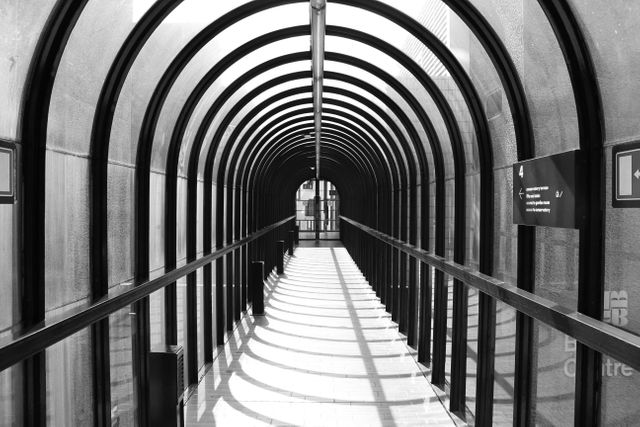 Monochrome perspective view of a modern glass walkway under harsh sunlight, creating strong shadows and linear patterns. Suitable for illustrating modern architecture, urban design concepts, or for use in design and art projects emphasizing linear perspective and abstract patterns.