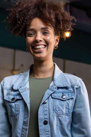 Smiling african american woman standing wearing denim jacket and olive top, gold hoops