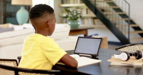 African american boy studying on tablet and writing notes at home living room workspace