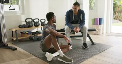 Diverse men chatting and stretching in home gym with kettlebells and bench