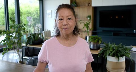 Senior asian woman standing in sunlit modern living room with lush houseplants and orchids