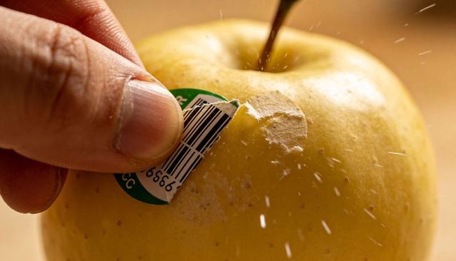 Peeling sticker from golden apple, hand removing barcode label, scattering adhesive bits closeup