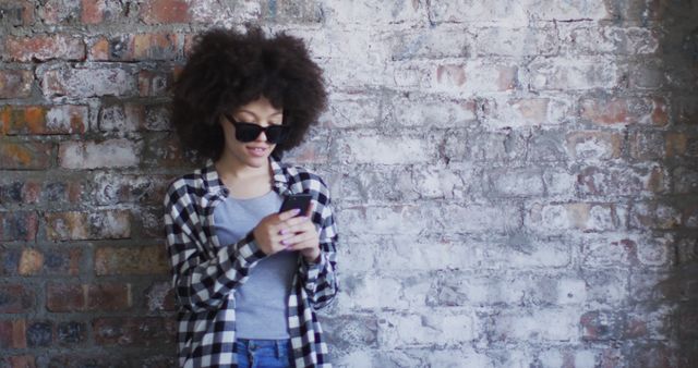African american woman wearing sunglasses using smartphone while standing against brick wall. concept of gen z gender expression identity and diversity.