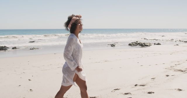 Person enjoys a stroll on the beach with clear blue sky overhead. White cover-up suggests casual and relaxed beach attire. Ideal for use in vacation guides, summer fashion, wellness blogs, and travel advertisements.