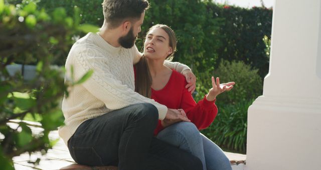 Young couple in knitted sweaters and jeans sits outside amidst lush greenery while having a heated discussion. Can be used in articles or campaigns about relationship issues, communication difficulties, or emotional expressions between partners.