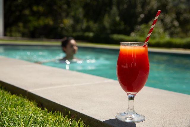 Refreshing cocktail with a red and white striped straw placed on poolside edge, with a woman swimming in the background. Ideal for use in summer vacation promotions, leisure and lifestyle blogs, travel advertisements, and outdoor relaxation concepts.