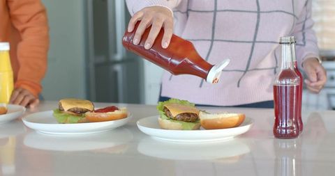 Couple assembling cheeseburgers pouring ketchup on buns in bright home kitchen