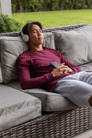 Asian man relaxing on rattan sofa listening to music with headphones on porch