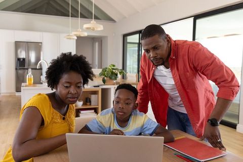 African american parents helping son with laptop homework at bright modern kitchen table