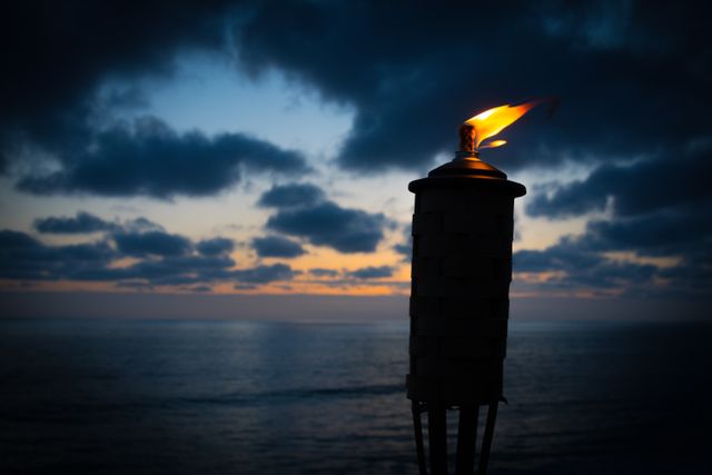 Tiki torch with flickering flame set against dark clouds and colorful sunset over calm ocean. Ideal for use in themes that evoke relaxation, tropical vacations, outdoor parties or serene beach settings.