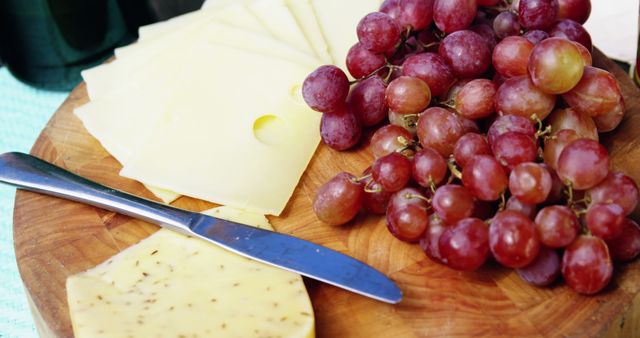 Assorted cheese and red grapes are on a round wooden platter. Slices of yellow cheese with small holes are neatly arranged next to a bunch of fresh red grapes. A wedge of seasoned cheese sits beside a butter knife. Ideal for illustrating gourmet food preparation, cheese boards, or wine and snack concepts. Useful for catering advertisements, food blogs, or social media content.