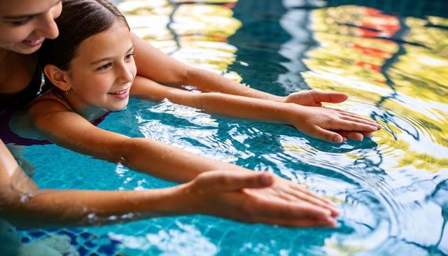 Young girl practicing glide while instructor guiding during swim lesson in sunlit pool