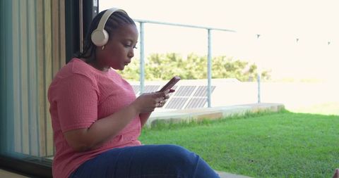Mid-30s african american woman relaxing on porch using smartphone and wireless headphones