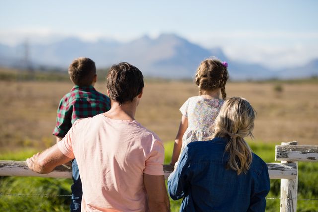 Family of four standing together, looking at scenic landscape with mountains in the background. Ideal for concepts of family bonding, outdoor activities, nature appreciation, and leisure time. Perfect for use in advertisements, brochures, and websites promoting family outings, nature parks, and outdoor recreation.