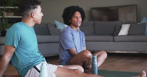 Two men sitting on yoga mats talking after home workout with water bottles in living room