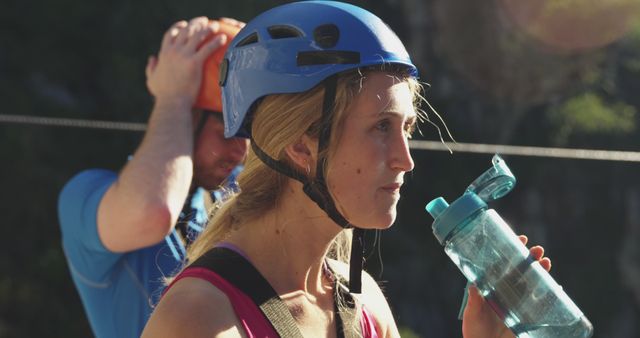 Young woman rock climber wearing a safety helmet, drinking water. Background showing another climber adjusting helmet. Ideal for content related to outdoor sports, climbing, fitness, adventure activities, and hydration tips during physical activity.