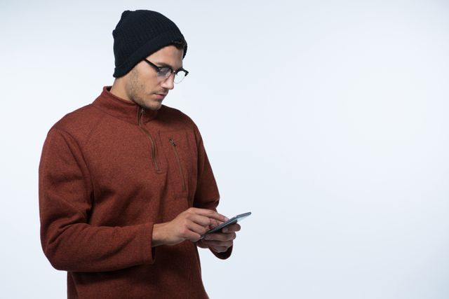 Young man wearing winter clothing and glasses, using a mobile phone against a white background. Ideal for themes related to technology, communication, winter fashion, and modern lifestyle. Suitable for advertisements, blog posts, and social media content focusing on tech-savvy individuals or winter apparel.
