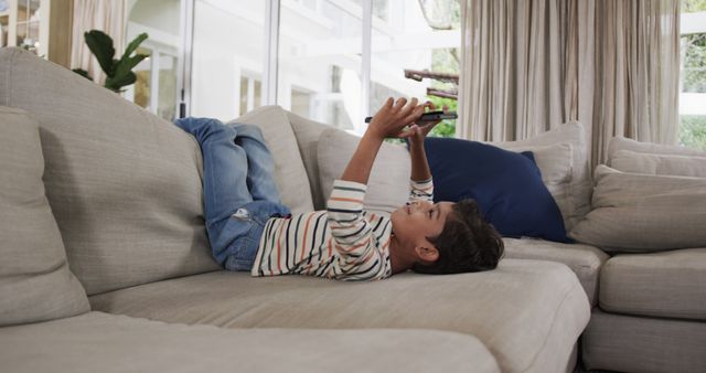 Young boy in casual clothes laying on back on a beige sofa engrossed in playing with a tablet. Background consists of a bright and airy living room with white curtains and window views of indoor plants. Ideal for illustrating technology use in children, modern living environments, or home leisure activities.