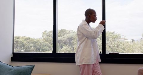 African clinician standing by window tasting spoon wearing lab coat and pink scrubs